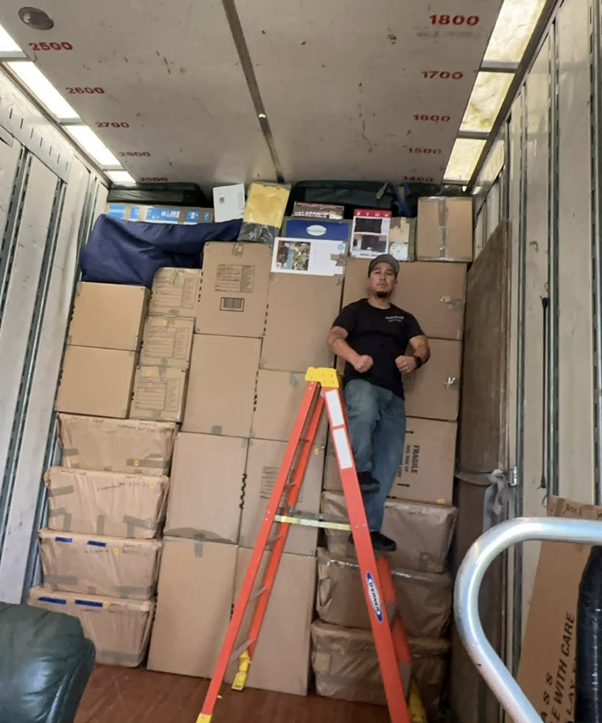 A mover standing atop a neatly stacked wall of packed boxes inside a truck